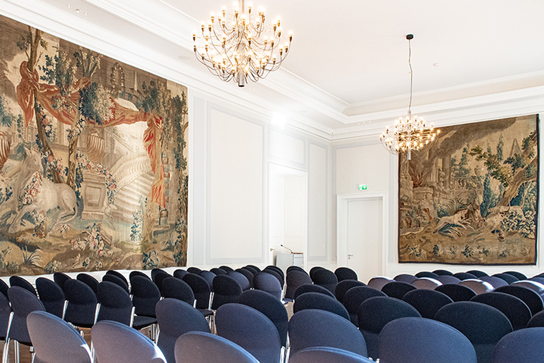 Blick auf den Gobelin-Saal des Alten Rathauses in Bonn, geknüpfte Wandteppiche, Kronleuchter an der Decke und blaue Stuhlreihen.