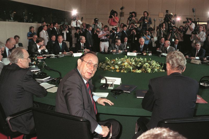 Round table with flower arrangements in the middle, in the fron Foreign Minister Hans-Dietrich Genscher, in the background many press representatives