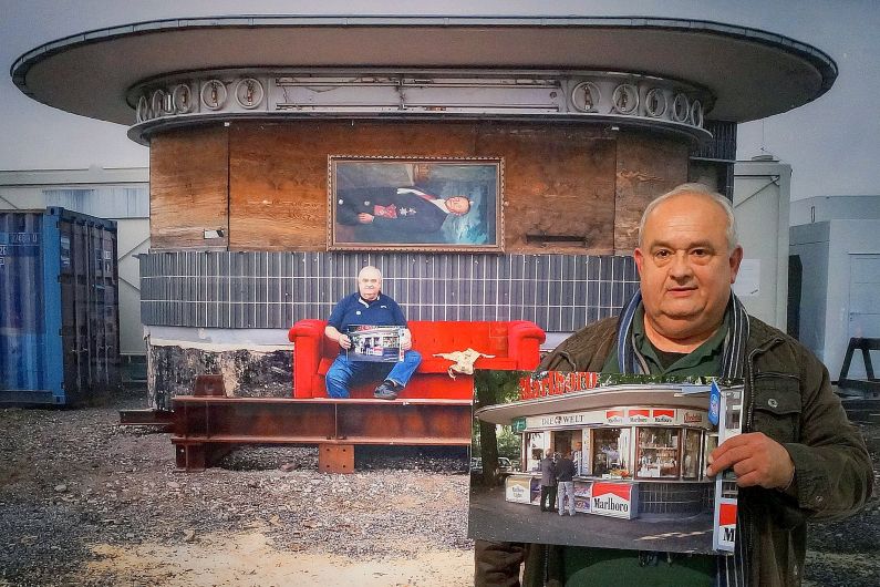 Ein älterer Mann hält ein Foto des Kiosks an seinem alten Standort hoch, im Hintergrund ist der Kiosk mit verbarrikadierten Fenstern auf einem Bauhof zu sehen.