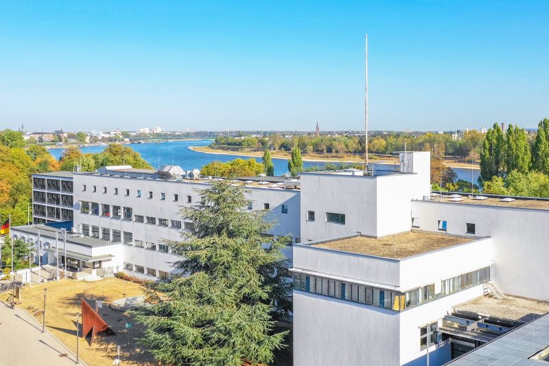 Luftaufnahme, weißer, kastenförmiger Gebäudekomplex, vierstöckig, Deutschland-Fahne am Eingang. Hinter dem Gebäude ist der Rhein zu sehen.