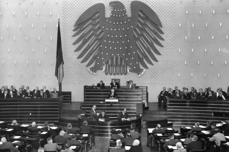 Schwarzweiß-Fotografie, vollbesetzter Sitzungssaal, mittig das Rednerpult, großer Bundesadler an der Wand, Deutschlandfahne links.