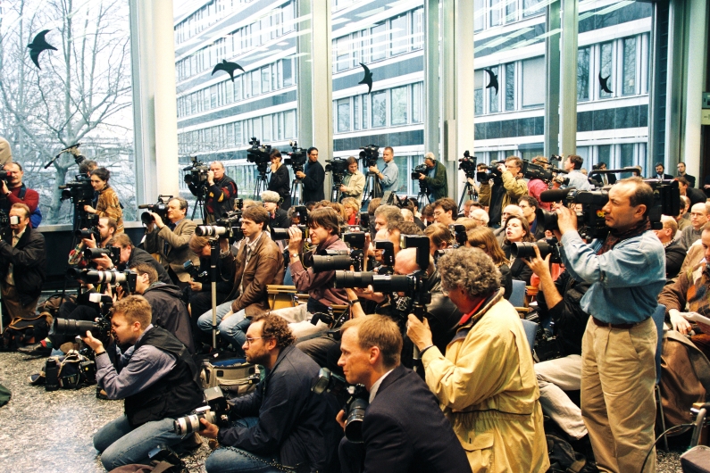 A large group of journalists in the press room of the Federal Press Conference in Bonn, sitting on several rows of chairs, equipped with numerous cameras.