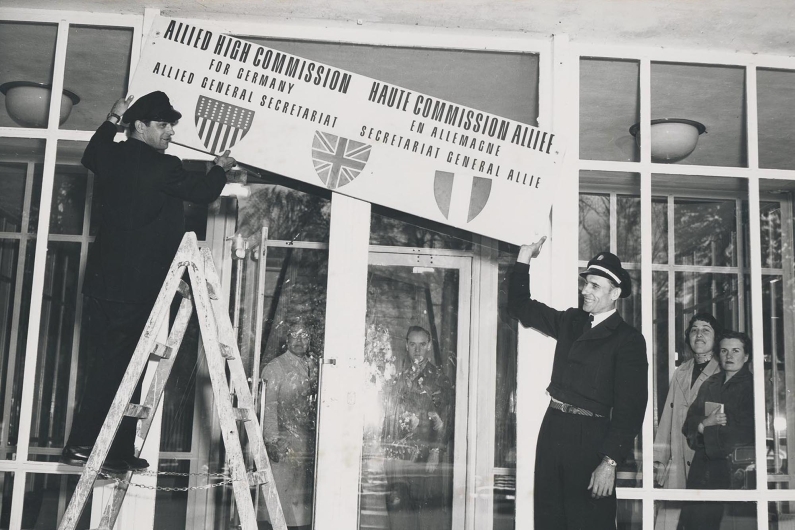 Black and white photograph, a man on a ladder removes the signage at the entrance of the US High Commission.