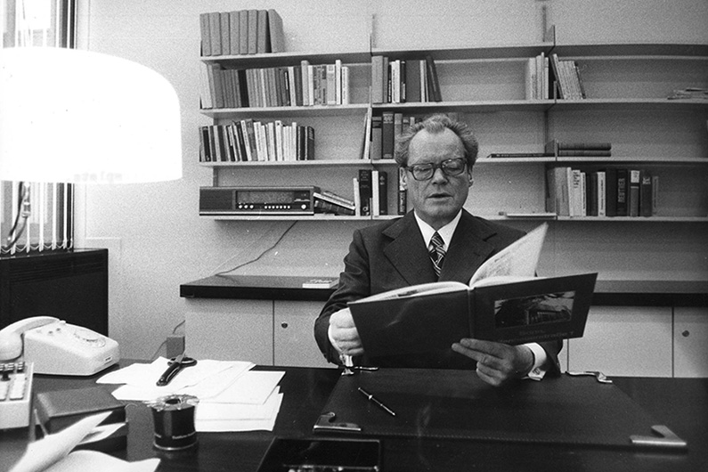 Black and white photography of Willy Brandt in his office, sitting at his desk with a book in his hand, behind him several shelves, on his desk a telephone and documents.