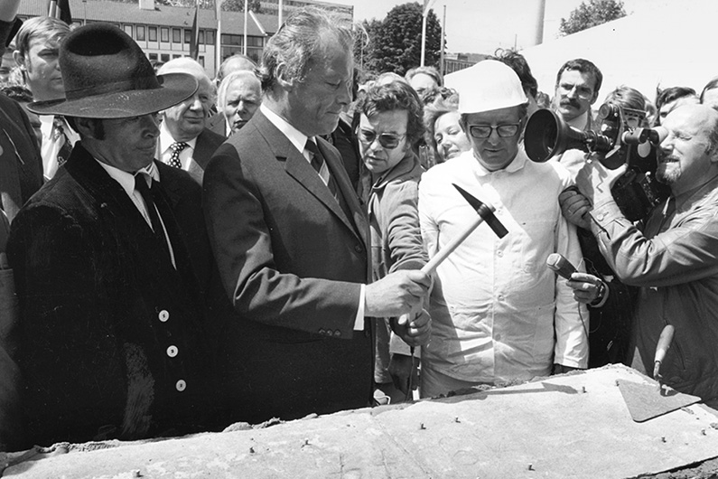 Schwarzweiß-Fotografie, Willy Brandt (mittig) bei der Grundsteinlegung des Erich-Ollenhauer-Hauses in Bonn, Pressevertreter filmen die symbolischen Hammerschläge Brandts mit dem Grundsteinhammer.