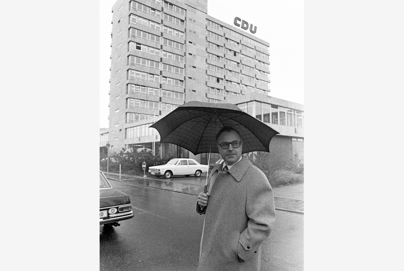 Schwarzweiß-Fotografie, Helmut Kohl steht mit Regenschirm vor dem Konrad-Adenauer-Haus in Bonn, links der zehngeschossige Plattenbau der CDU-Parteizentrale, davor die "Union-Säle" in einem Flachbau.