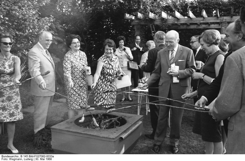  Several people around a barbecue grill where they grill sausages on sticks.