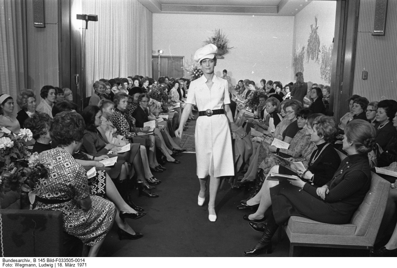 A model in the middle of a catwalk surrounded by a female audience. The model is wearing a white outfit with a hat