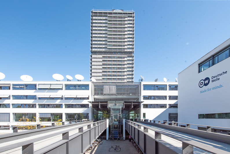 A paved path leads frontally to the entrance of Germany’s international broadcasting service "Deutsche Welle", behind of it you can see the "Langer Eugen" office tower.