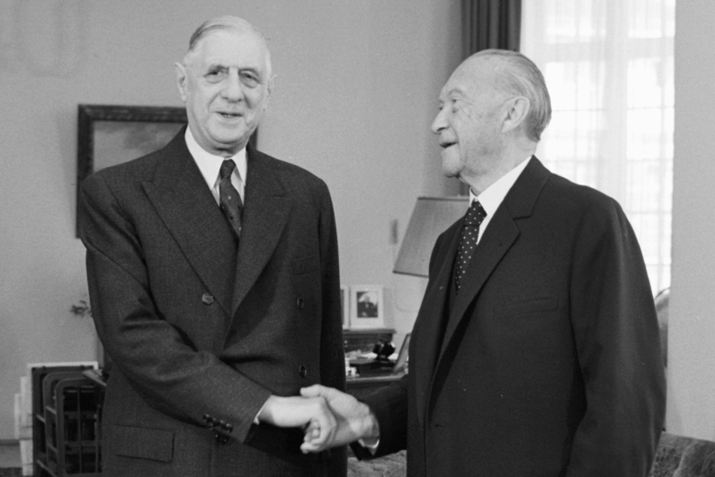 Konrad Adenauer and Charles de Gaulle in suits, standing in Adenauer's study and shaking hands.