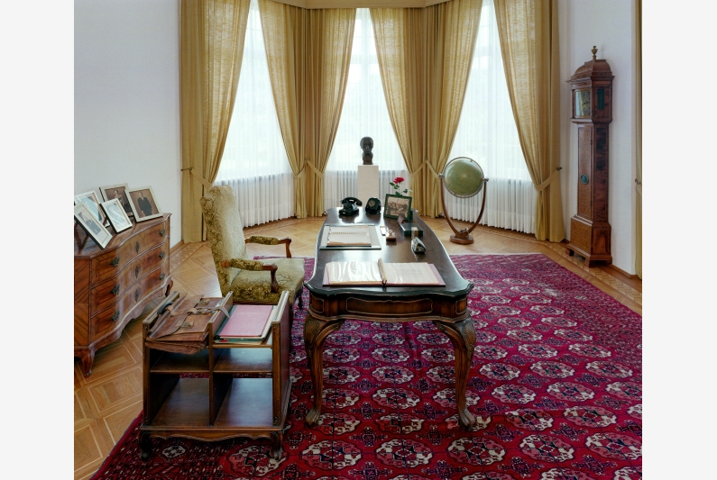 Study with desk and wooden furniture, a red woven carpet and large bay windows.