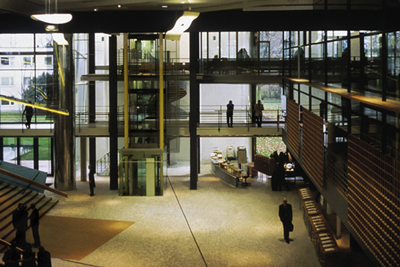 View into the generous entrance area of the Bundeshaus, large glass windows, light-coloured floor, a glass elevator in the middle, a wide wooden staircase to the right of it.