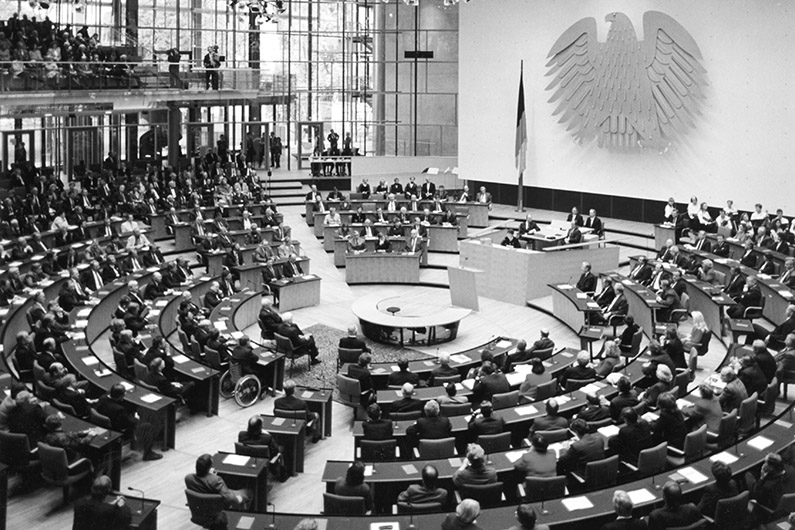 Schwarzweiß-Fotografie, Blick auf einen gefüllten Plenarsaal mit kreisrunder Sitzanordnung, heller und transparenter Architektur, einem Rednerpult, an der Wand dahinter der Bundesadler