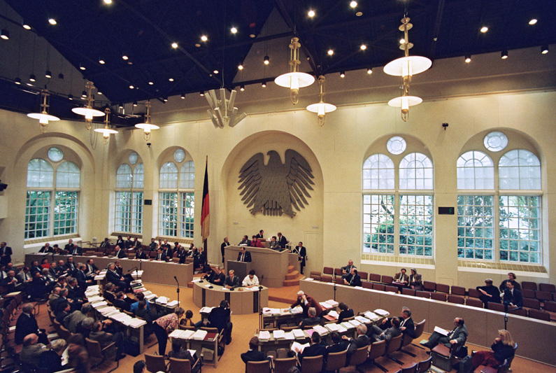 Blick auf den ersten gesamtdeutschen Bundestag im Plenarsaal des Wasserwerks in Bonn, die Parlamentarier in kreisrunder Sitzanordnung, mittig das Rednerpult, dahinter eine Wand mit großem Bundesadler und Schmuckfenstern