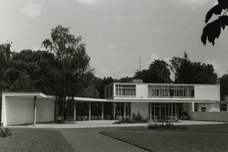 Schwarzweiß-Fotografie eines Pavillon-ähnlichen Teils der Stadthalle mit Gartenstühlen und Rasen davor.