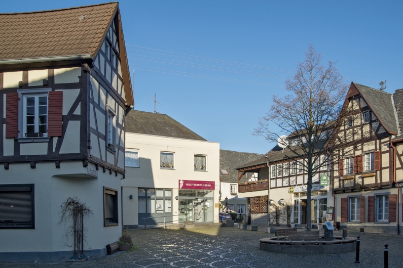 Unkeler Marktplatz mit Kopfsteinpflaster bei blauem Himmel, hinten mittig das Willy-Brandt-Forum, an den Bildrändern Fachwerkhäuser, vorne rechts ein Baum